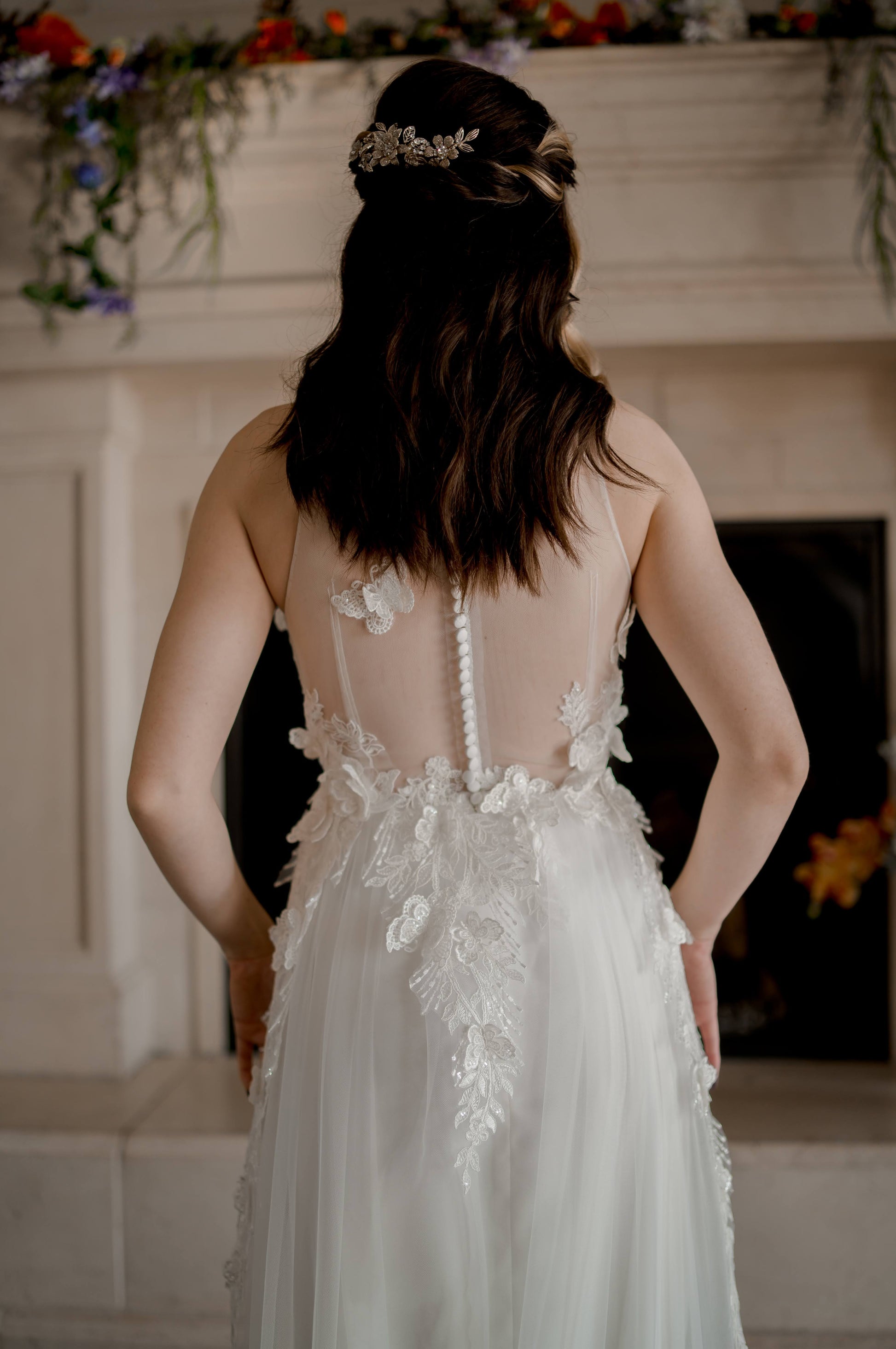 A bride stands in front of the fire place wearing the jacqueline gown. She faces away showing off the  Illusion back with satin buttons and scattered lace butterflies.