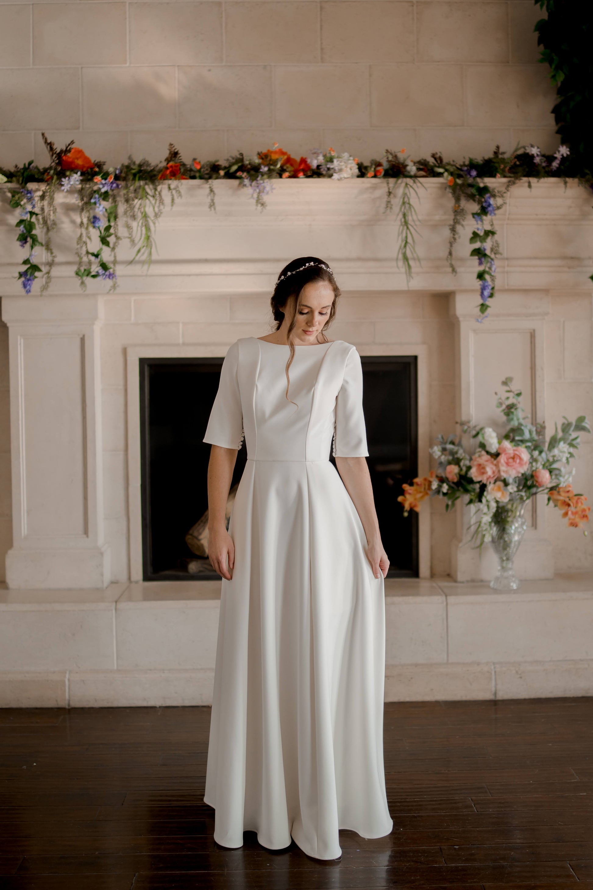 A full length image of the Wendy gown; a bride stands in front of a fire place with the boat neck, elbow length sleeves and a-line silhouette on display.