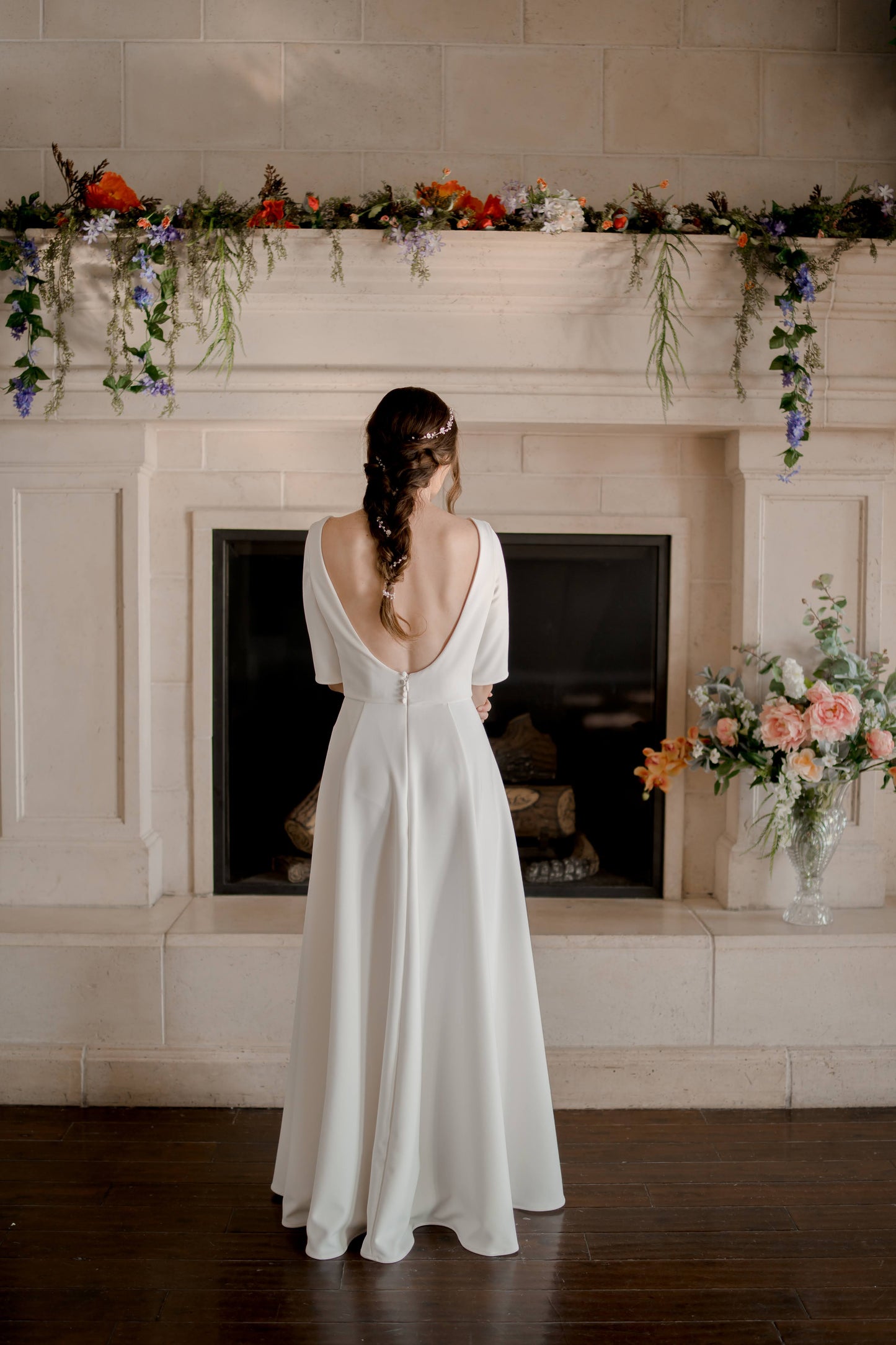 A full length back shot of the wendy gown; the bride stands in front of a fire plac with the low scooping back and silhouette on display. the gown does not have a train.