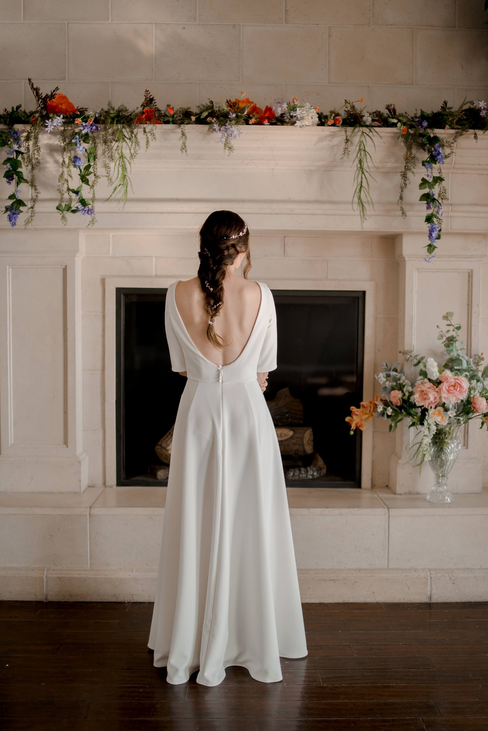 A full length back shot of the wendy gown; the bride stands in front of a fire plac with the low scooping back and silhouette on display. the gown does not have a train.
