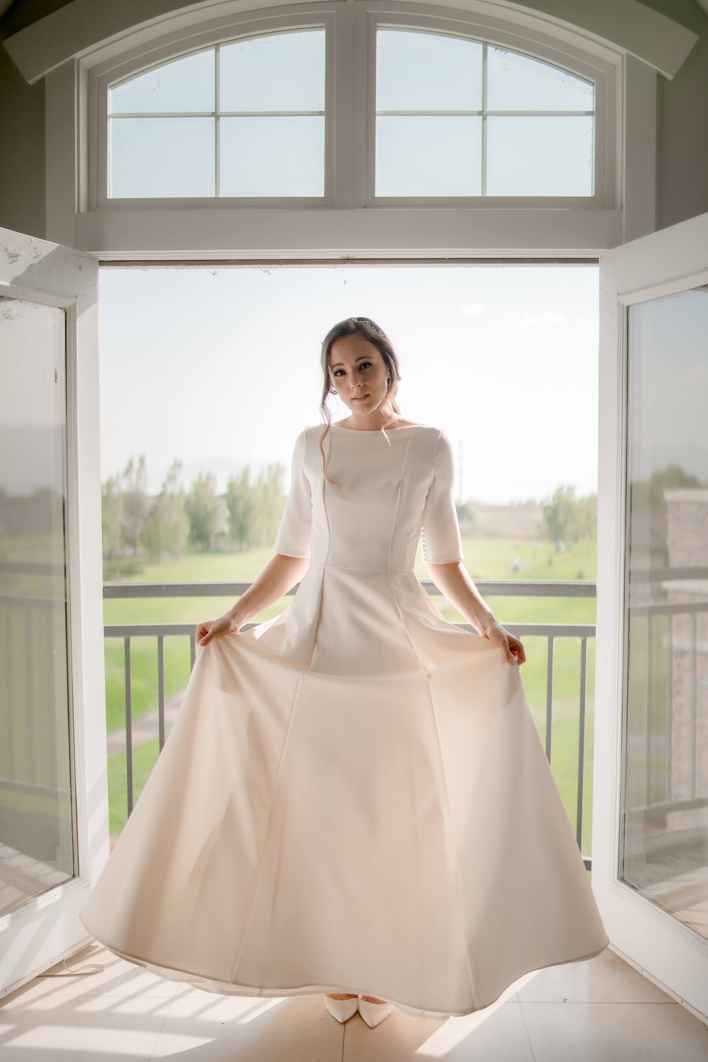 A full length photo from the front of a bride inbetween a set of double doors; she holds the a-line crepe skirt out to it's full widh showing off the skirt and structured bodice.