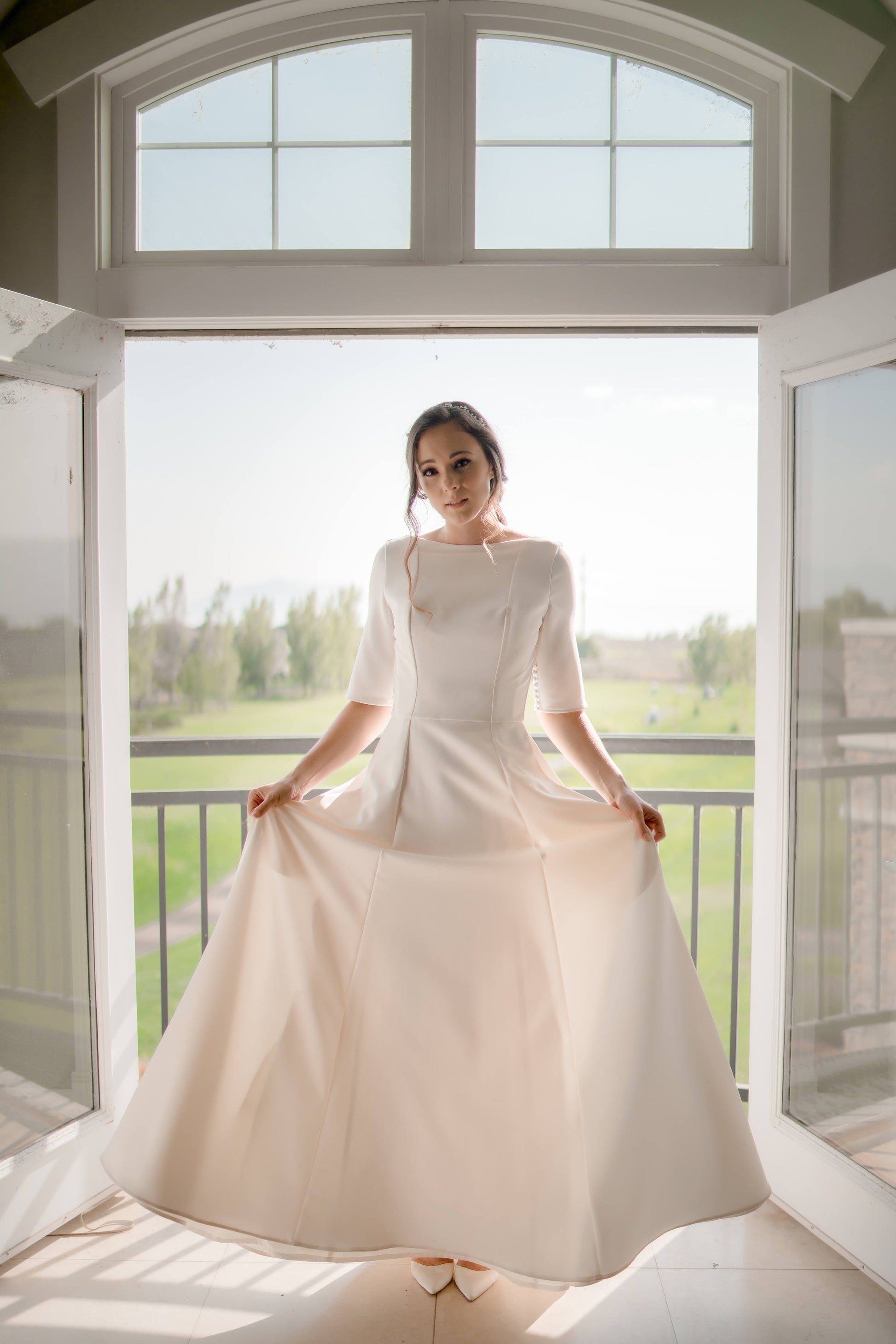 A full length photo from the front of a bride inbetween a set of double doors; she holds the a-line crepe skirt out to it's full widh showing off the skirt and structured bodice.