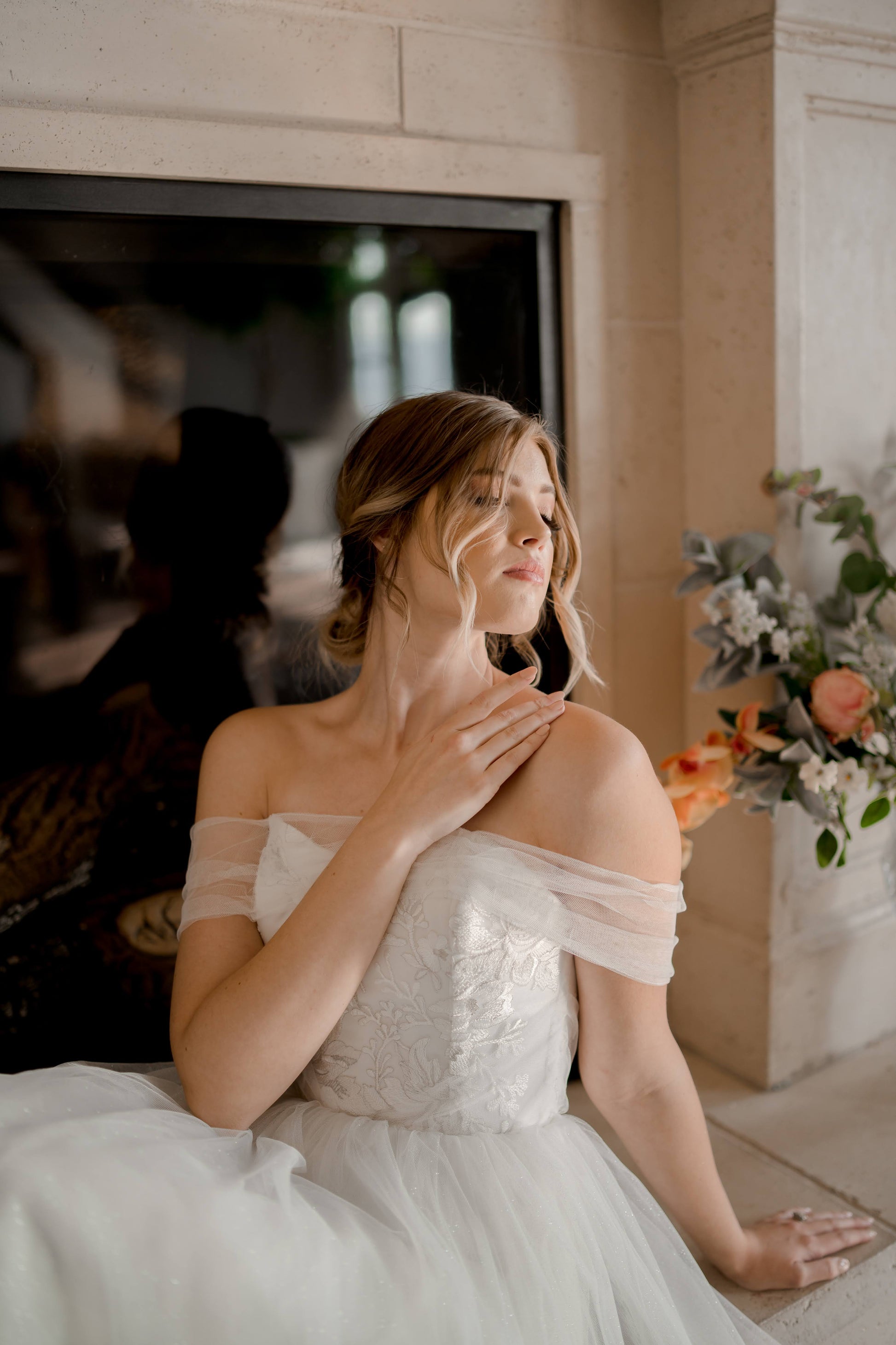 A styled shot of a bride wearing the Penelope gown, this focuses on the bodice and upper skirt as the bride sits on a hearth. The off the shoulder arm drape, and tulle skirt are prominent.
