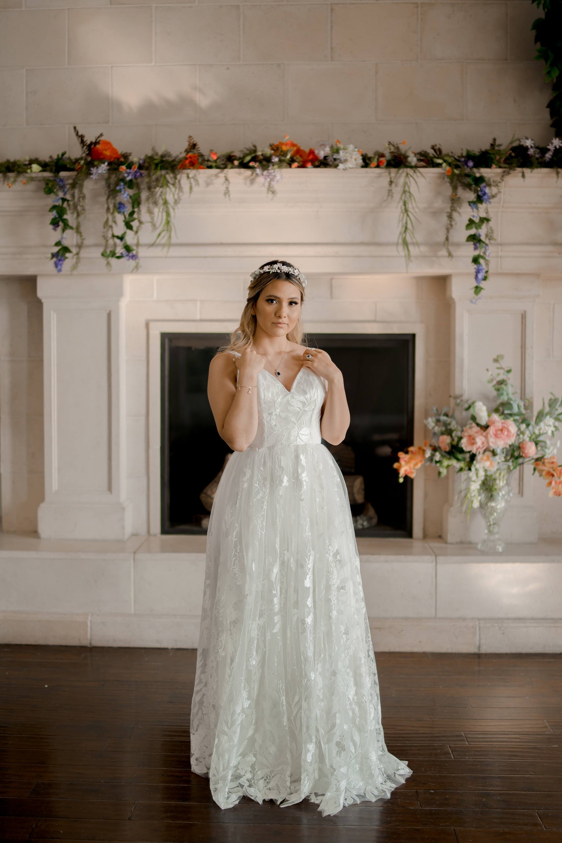 A brides wears the Rosie gown in standing front of a fireplace; the wedding dress has a gathered lace skirt, v neckline, and a modified a-line silhouette.