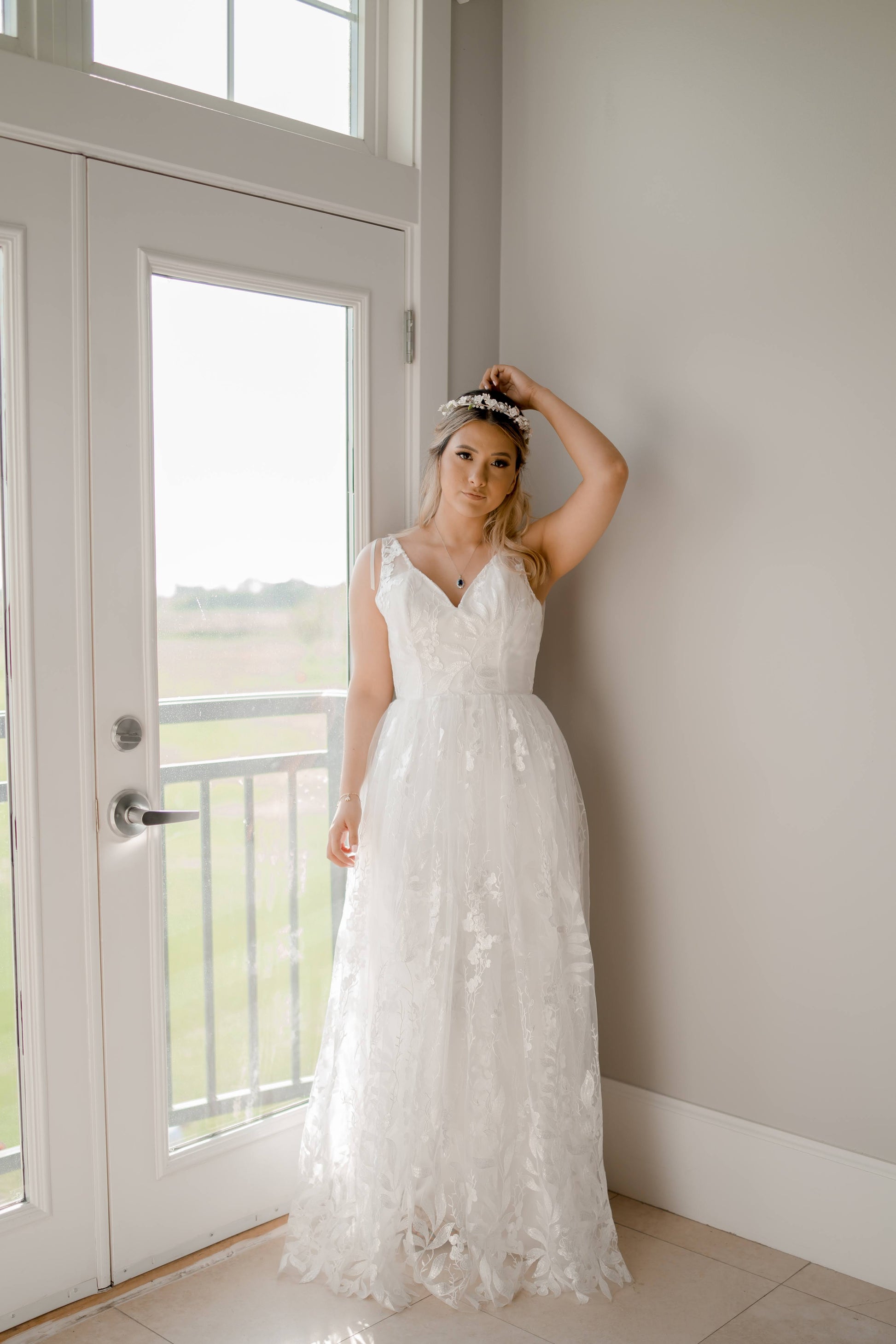 A styled shot of a bride wearing the Rosie wedding gown; she stands near a glass door and the gathered lace skirt, with satin sweetheart neckline, and tank top style straps are the focus.