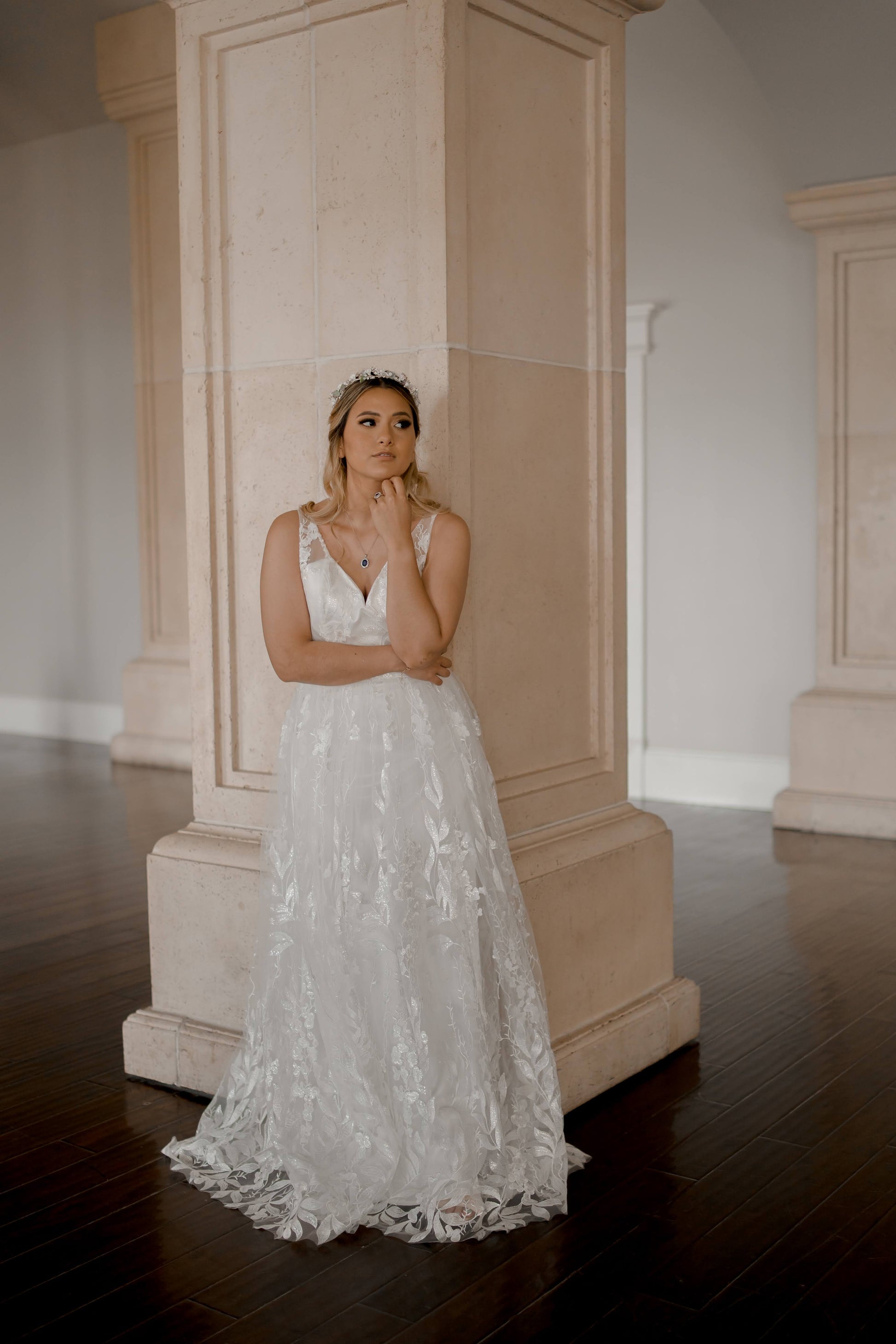 A styled shot of the Rosie gown on a bride; we can see the full front of the lace covered modified a-line gown as she leans against a pilar.