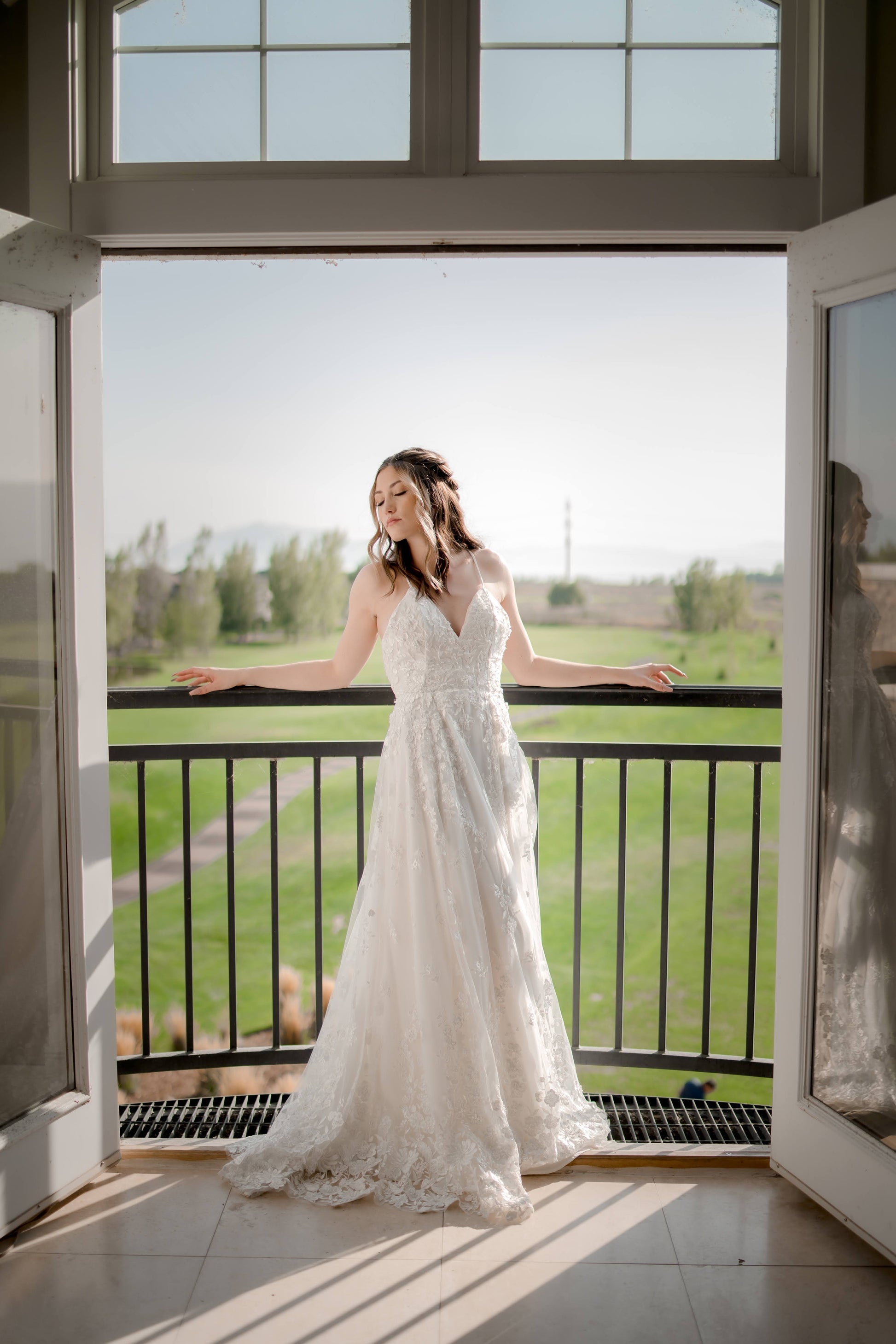 A full length shot from the front of a bride showing off the drape and fullness in the skirt of the Wilhelmina gown. She leans on a balcony railing between double doors.