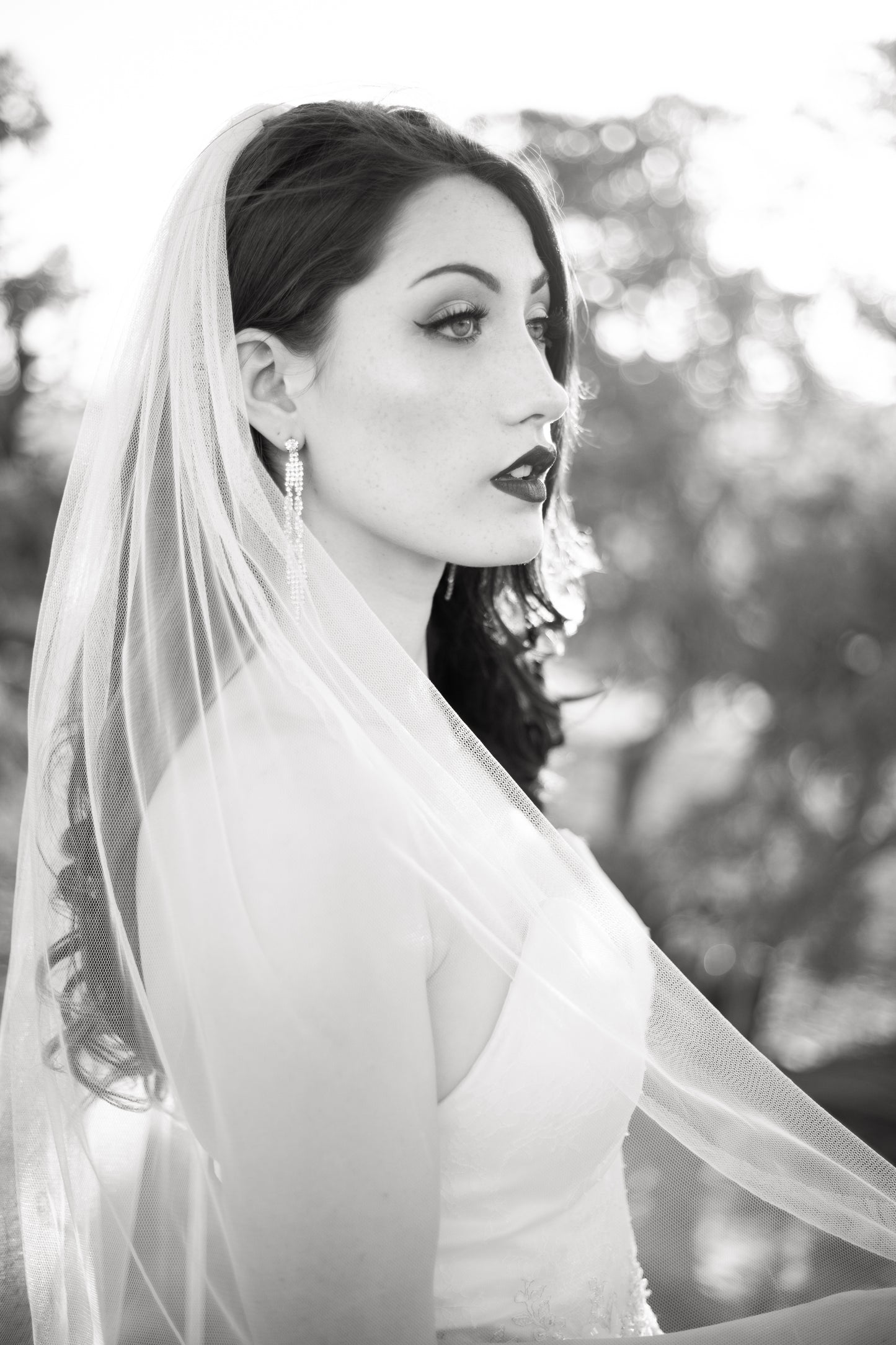 A bride wears the minimalist bridal veil, Jane. The bride pulls the edge of the veil forward as it falls from her hair. She stands in profile and looks off into the distance. 