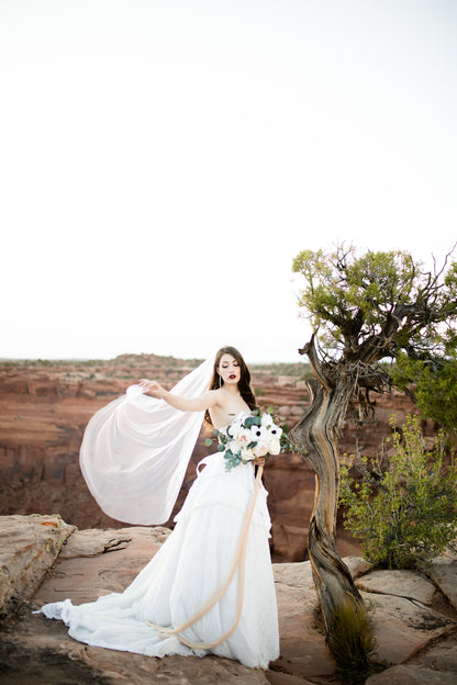 A full length photo of a bride in the desert wearing a layered ball gown and the waltz length veil Jane. The veil is mid air as the bride holds the tip and it is caught in the breeze. 