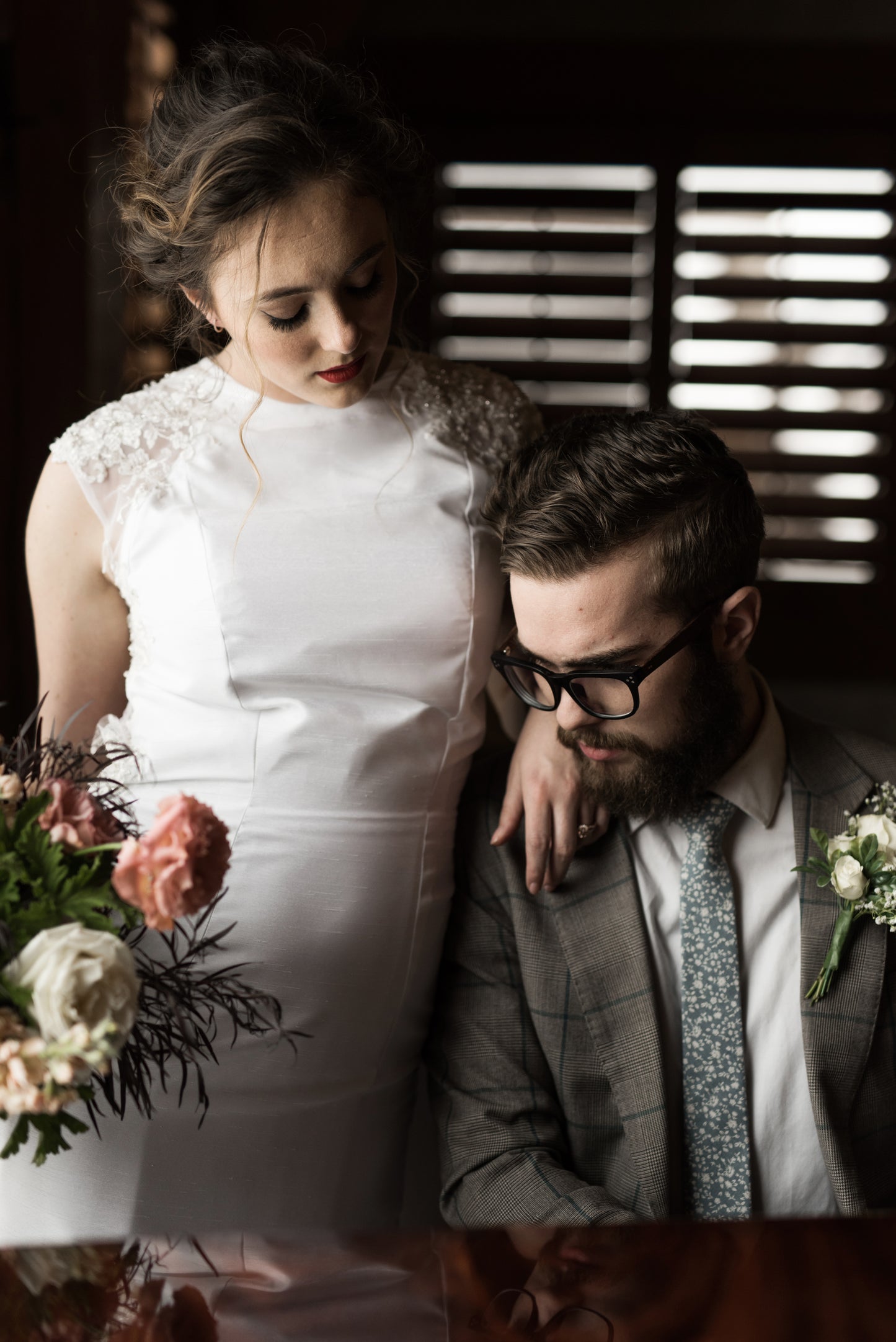 A bride leans on her groom while he plays piano, she wears the Mia gown; a Shantung halter neckline with darted bodice and beaded lace shoulders. 