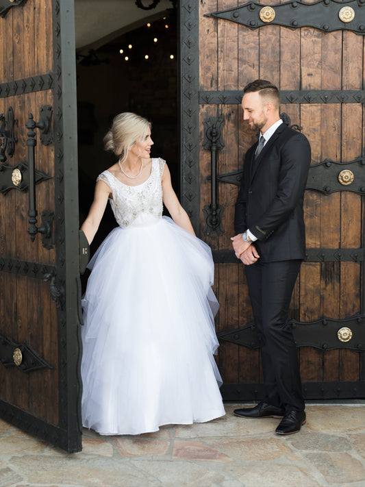 A bride steps through a castle door showing the front view of the Georgia wedding dress with scoop neckline, rhinestone bodice, and tiered tulle skirt.