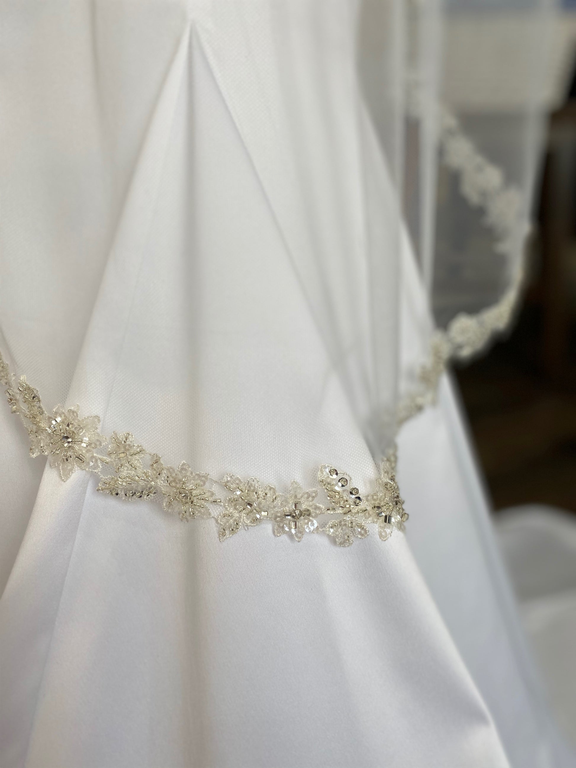 A side close up of the hem of the Glam bridal veil with lace and sequin edge, Haven. The gown is laying against a satin mermaid gown. 