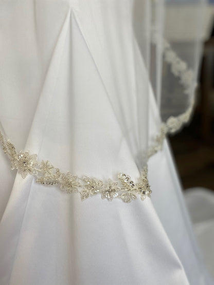 A side close up of the hem of the Glam bridal veil with lace and sequin edge, Haven. The gown is laying against a satin mermaid gown. 