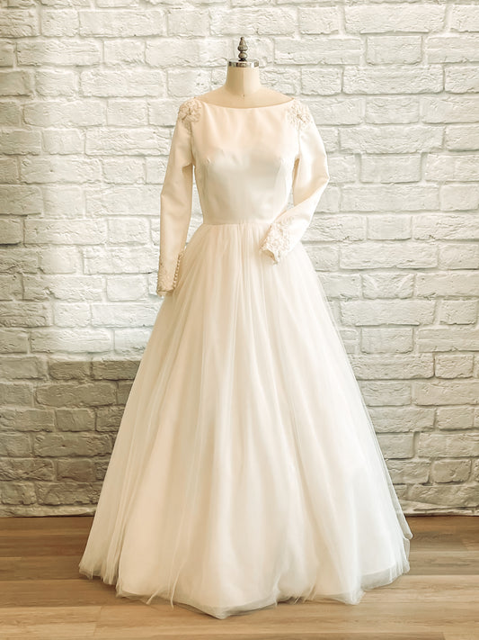 mannequin wearing the Ava gown with a boat neckline, long satin sleeves, and a full soft tulle skirt, standing gracefully in soft light.