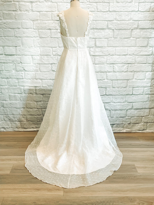 Back view of the Aunna wedding dress, showing the straight neckline, floral straps trailing down the back, and layered train of organza extending gently behind.