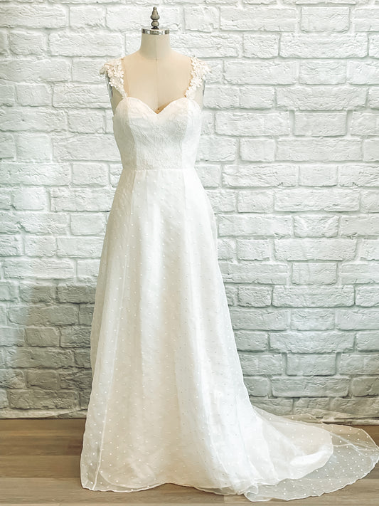 A mannequin wearing the Aunna gown facing forward, showcasing the romantic A-line silhouette with layered embroidered organza and a sweetheart neckline framed by delicate 3D floral straps.