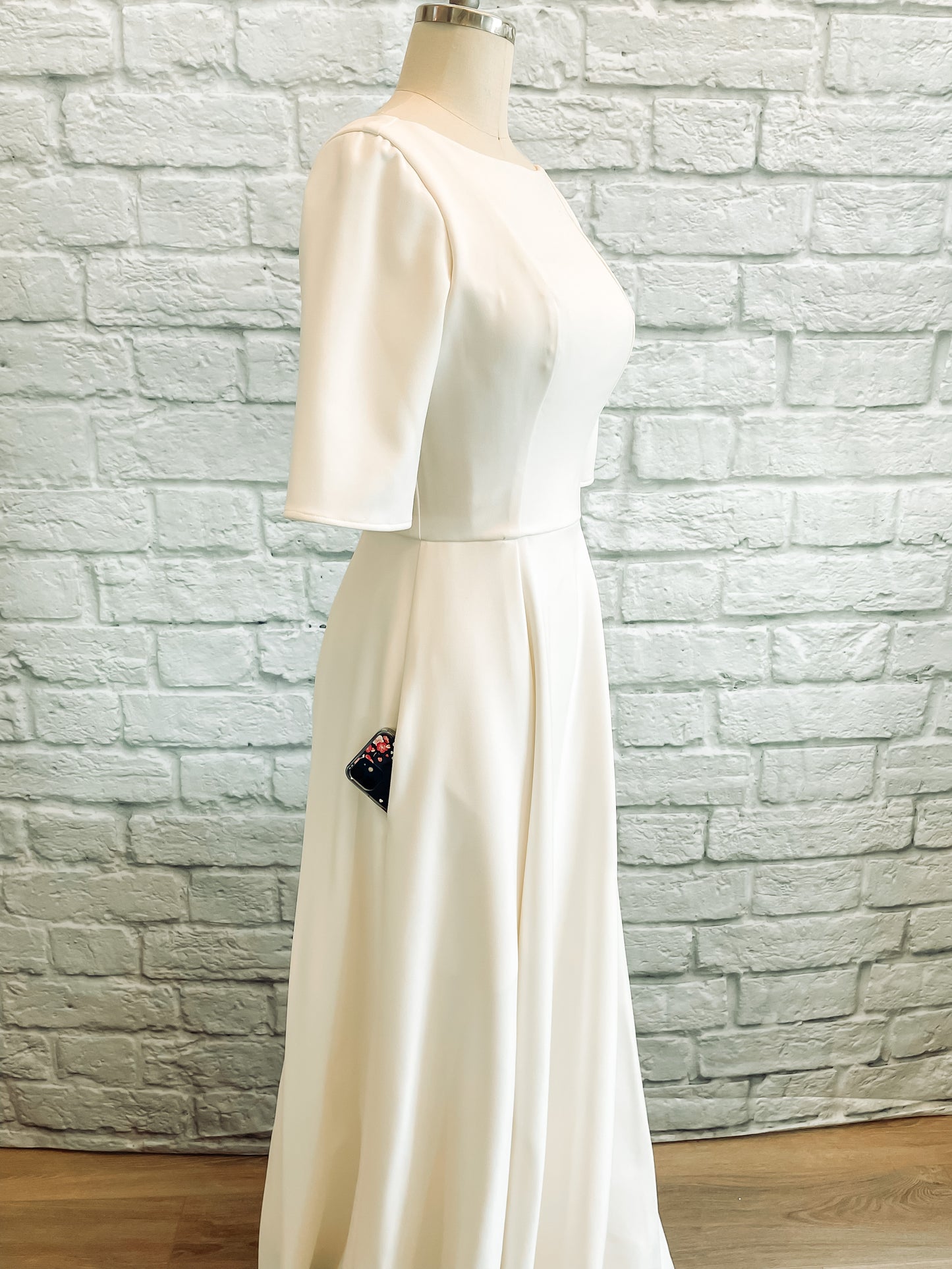 A profile view of the wendy gown on a mannequin; with the seam-matched skirt fullness, smooth stretch crepe fabric, and hidden pockets of which a phone pokes out are all featured. 