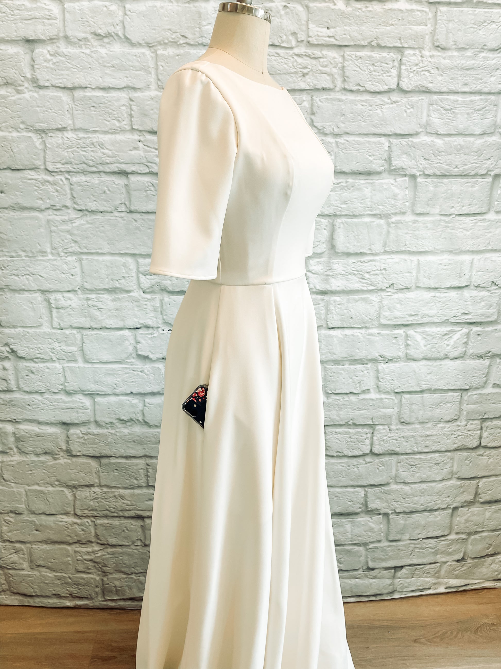 A profile view of the wendy gown on a mannequin; with the seam-matched skirt fullness, smooth stretch crepe fabric, and hidden pockets of which a phone pokes out are all featured. 