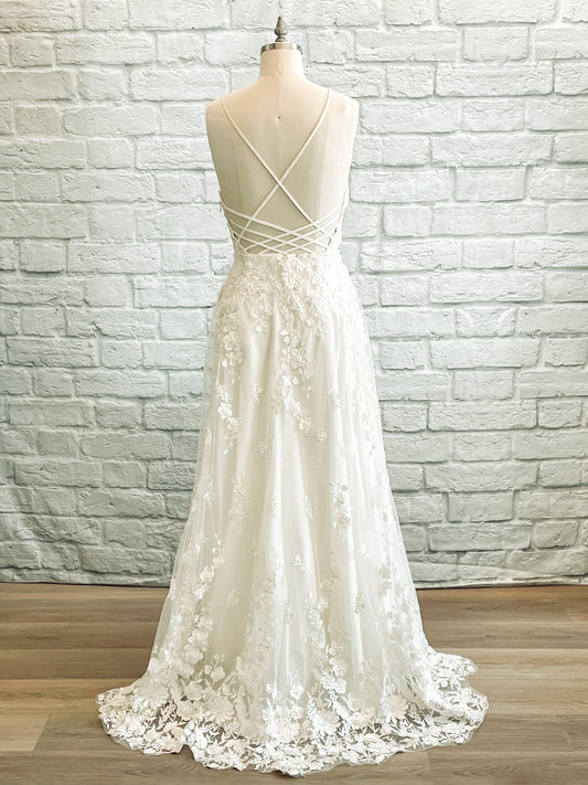 A full length shot of the wilhelmina gown from the back; the gown has  Crisscrossing straps over an open back, lace skirt, and small lace train detail