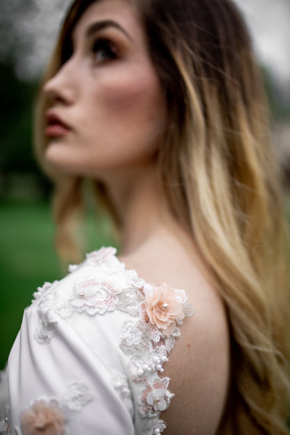An upclose photo of the lydia gown's shoulder on a bride, highlighting the 3D floral lace with pink threading, pearls, and sculpted petals. 