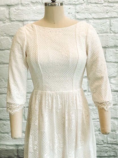 Detail shot of Avabelle gown’s front bodice , lace sleeves and wide boat neckline on a mannequin, highlighting the rose lace pattern and scalloped eyelash edges.