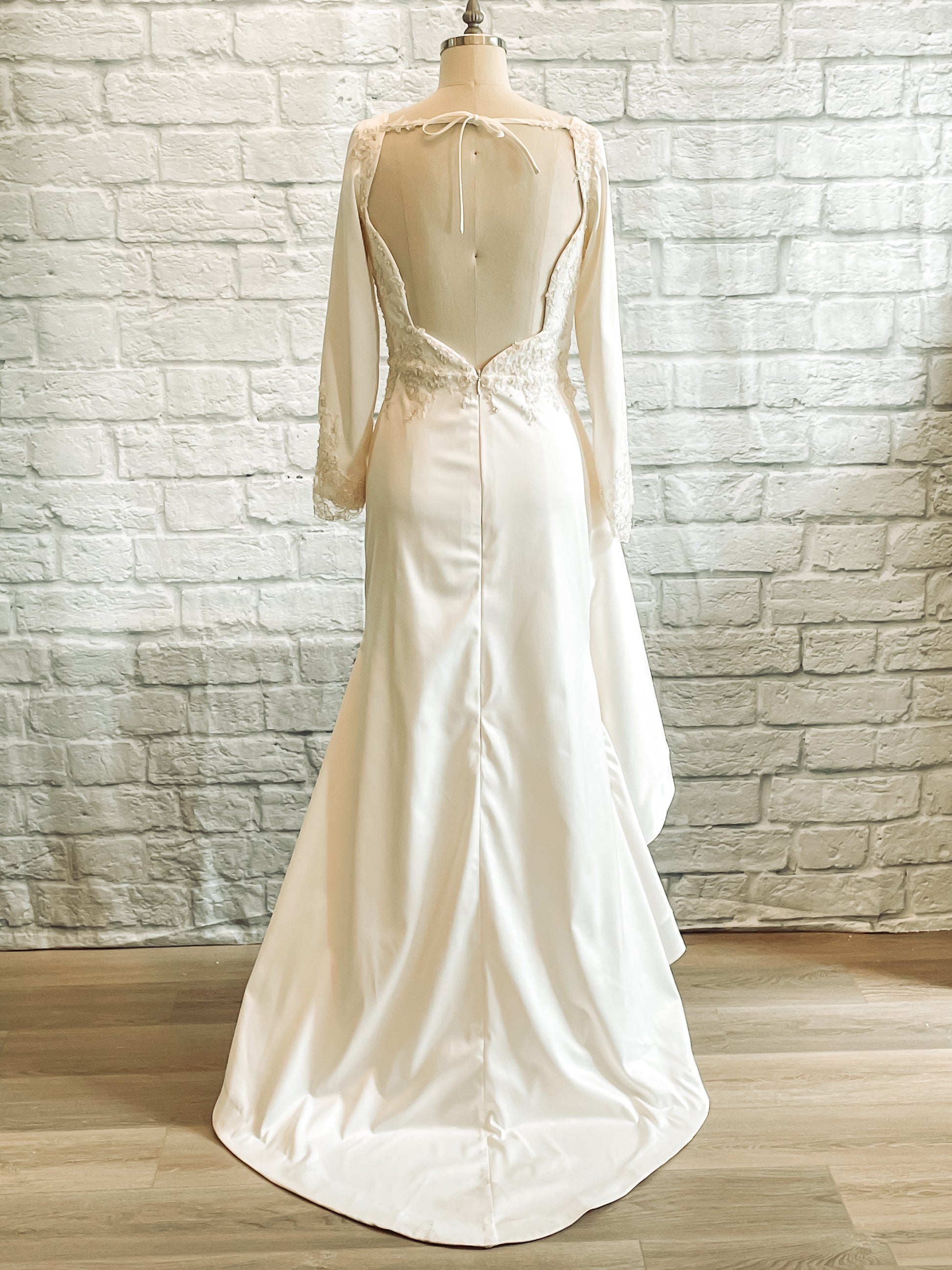 Rear view of Emma gown on mannequin, with architectural back neckline, long lace-trimmed sleeves, and small train. 