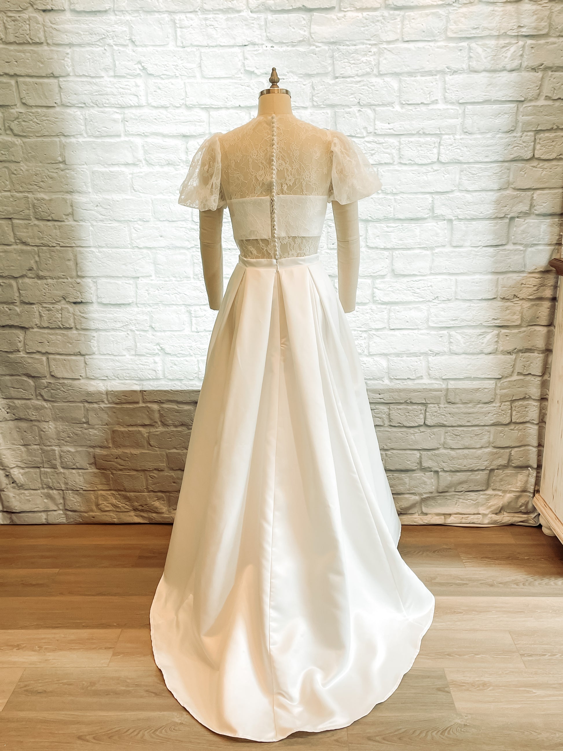 Back veiw of the Jessa gown; Lace back with satin-covered bridal buttons on sheer and bando layers, pleated satin skirt, and small train. 