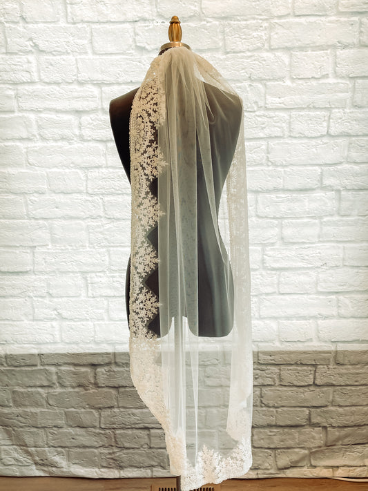 A calf length photo of the Ireland veil with Medium-weight tullé, scalloped trim and floral accents. the veil is displayed on a mannequin with one edge layed over the shoulder. 