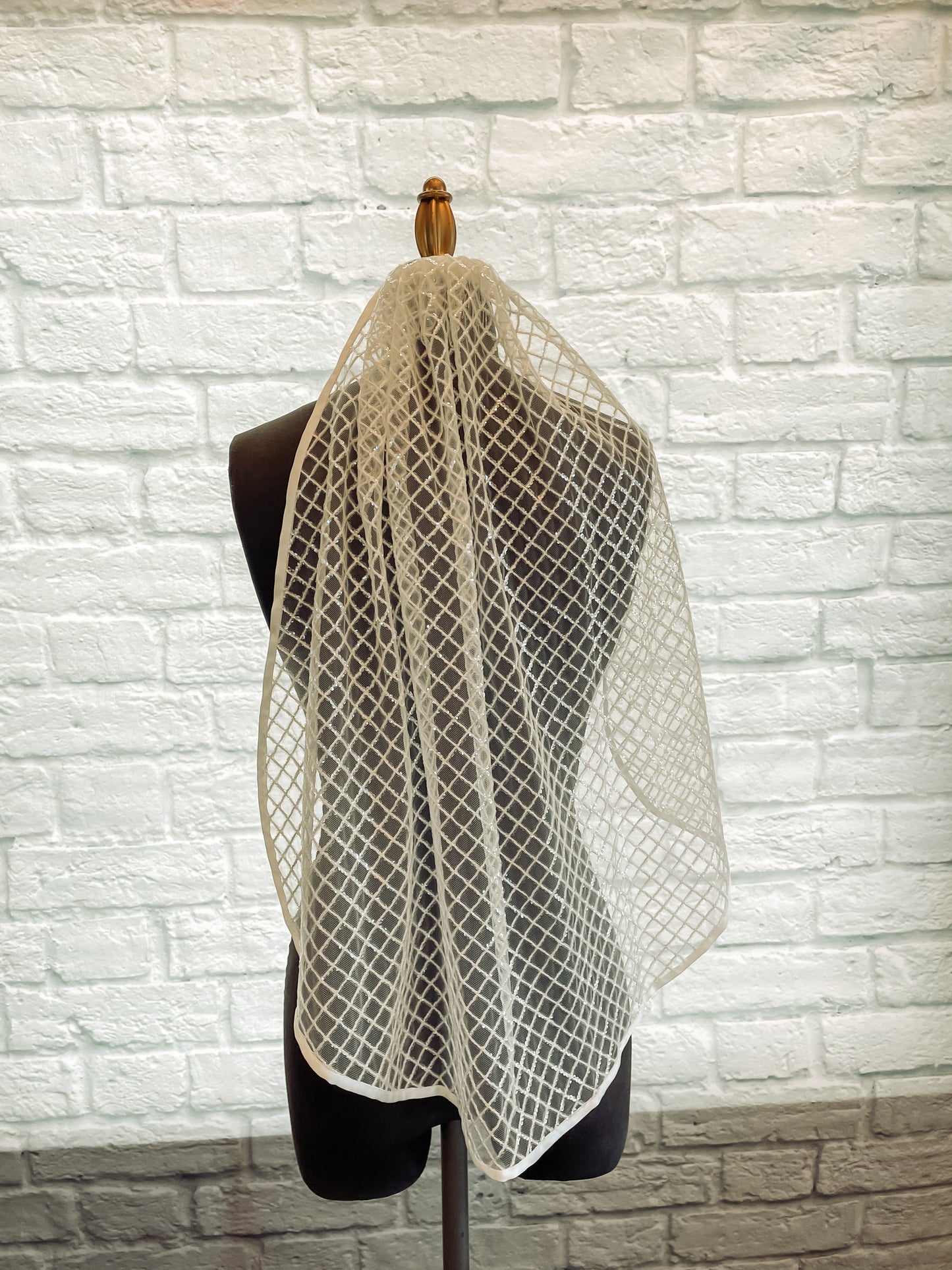 The gloria veil has been displayed on a mannequin with the veil pulled over one of the shoulders and not the other. This veil is covered in sequins in a diamond pattern and ribbon trim edge creating a detailed fairytale look. 