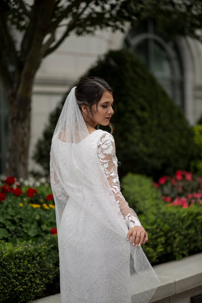 in this photo the scarlett veil is being paired with the octavia gown. a bride stands with her back to us and holds the edge of the swiss dot veil out behind her. The waltz-length veil with playful Swiss Dot texture pairs with the all lace gown octavia. 