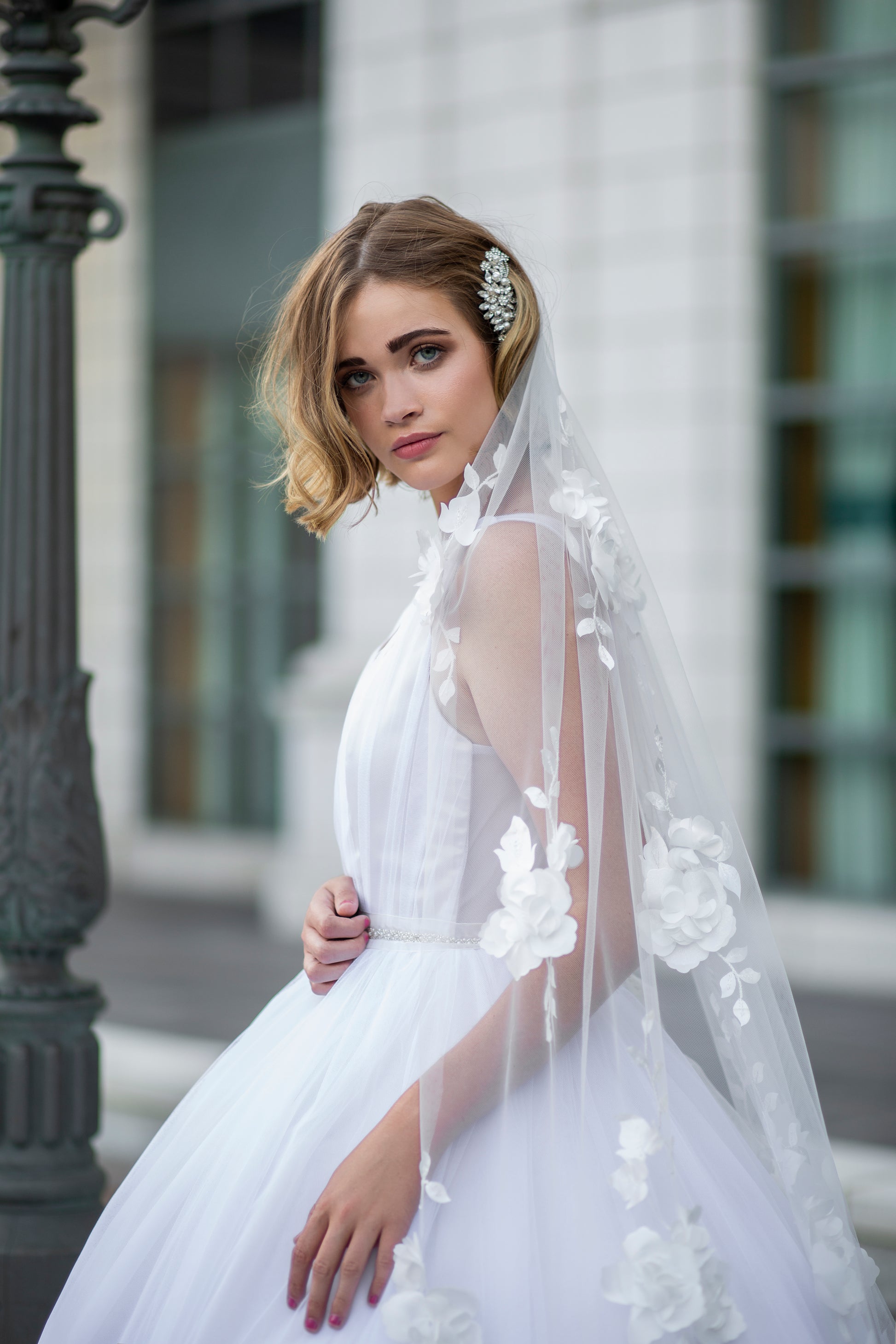 A photo of a bride wearing the persephone gown, fleur belt and celine veil. the image is taken from profile witht he bride looking at the camera. The veil has been swept to the side over the brides shoulder with the floral lace wedding veil the focus. 