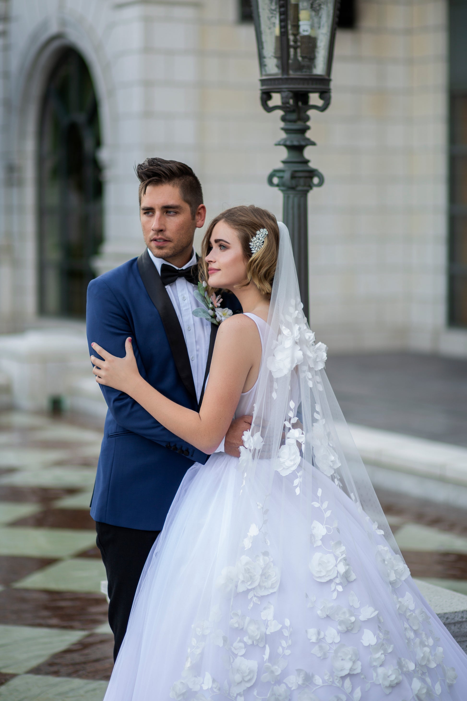 A bride and groom stand together embrasing. She wears the persephone gown and Celine veil. The veil is the focal point of the image as it lays over the ball gown showing off the 3d flowers. 
