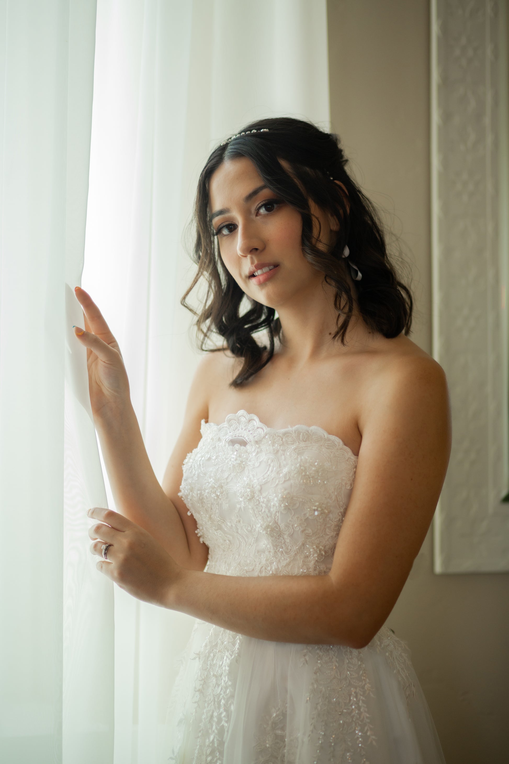 Model stands near a window with curtians wearing the kennsington tulle a-line gown; the beaded bodice, scalloped neckline and tulle skirt are all in focus. 