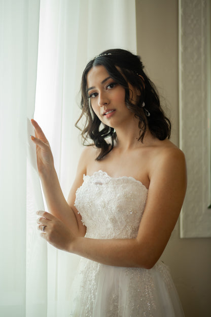 Model stands near a window with curtians wearing the kennsington tulle a-line gown; the beaded bodice, scalloped neckline and tulle skirt are all in focus. 