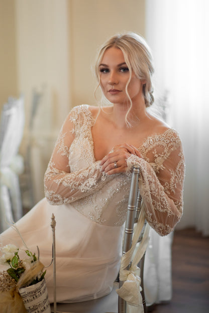 Bride sits sideways on a chair and leans  with her arm over the back of the chair. The sleeves and lace covered bodice are shown off, along with the tiny waist created with a defined seam  . 