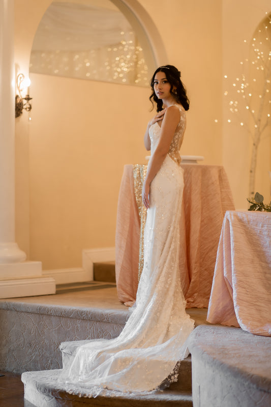 A side profile of the lorien gown showing the Sheath silhouette, fitted bodice, lace overlay detail, all with the train swept to the side, trailing down some stairs. 