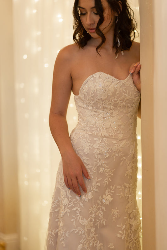 A ¾ view of the Sawyer wedding gown; A model wears the gown and leans against the wall. The trumpet silhouette is showcases the curve-accentuating fit and lace placement. 