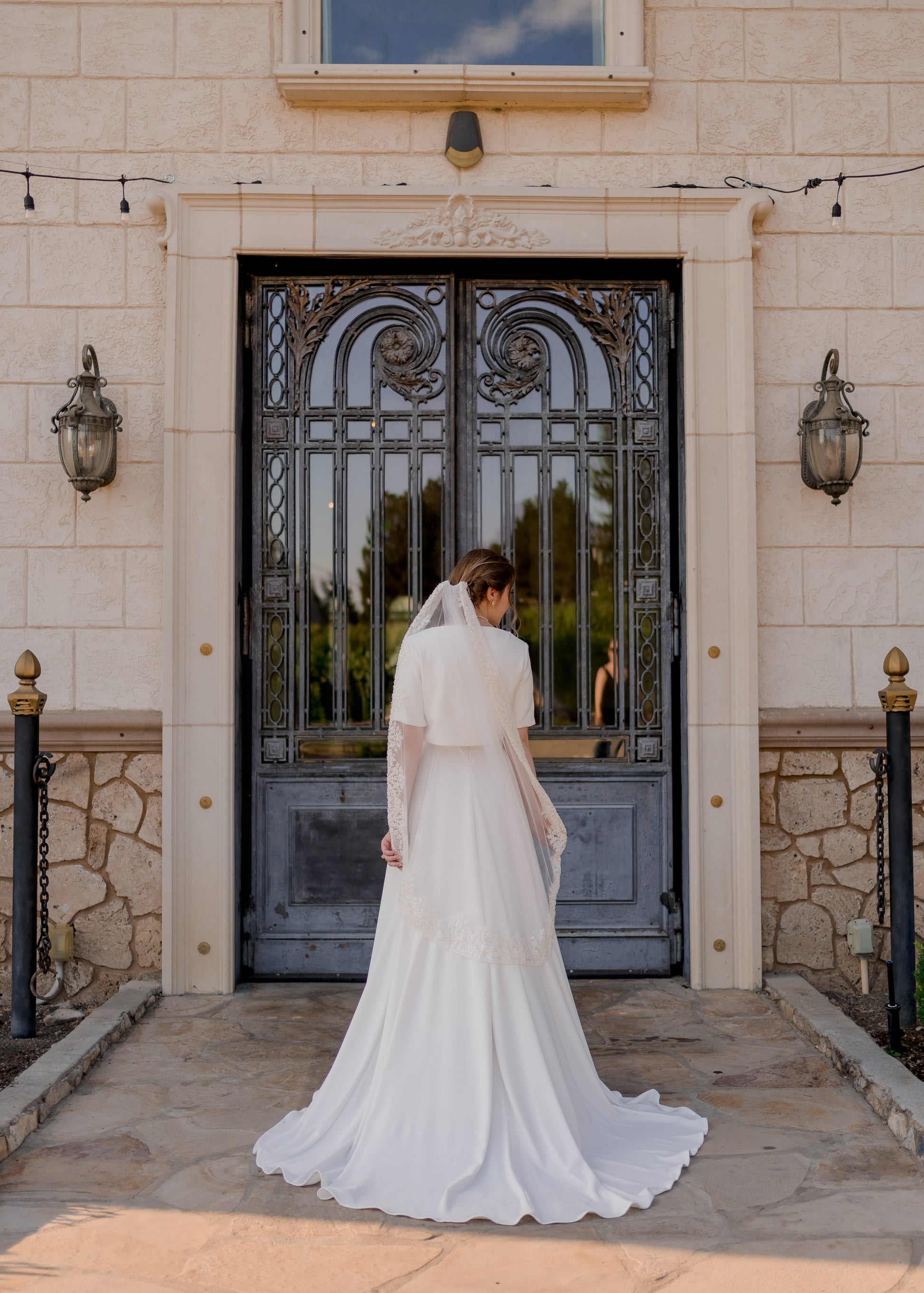 A full length back shot of a bride wearing the Cassie set and Ireland beaded veil. The bride holds the edges of the veil with her fingertips. The veil is in a pale ivory color and in a waltz length. 