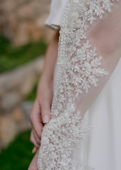 A close up of the breaded trim edge of the Ireland veil over a brides arm. the bride is wearing the crepe cassie set; you can see behind the veil and brides arm. 