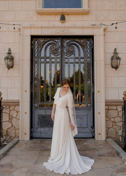 A photo of a bride; she stands to the side and leans down to the side and slightly back with her arm stretching out in the same direction. She wears the crepe bridal set, Cassie and the beaded trimmed bridal veil, Ireland, in a calf length. 