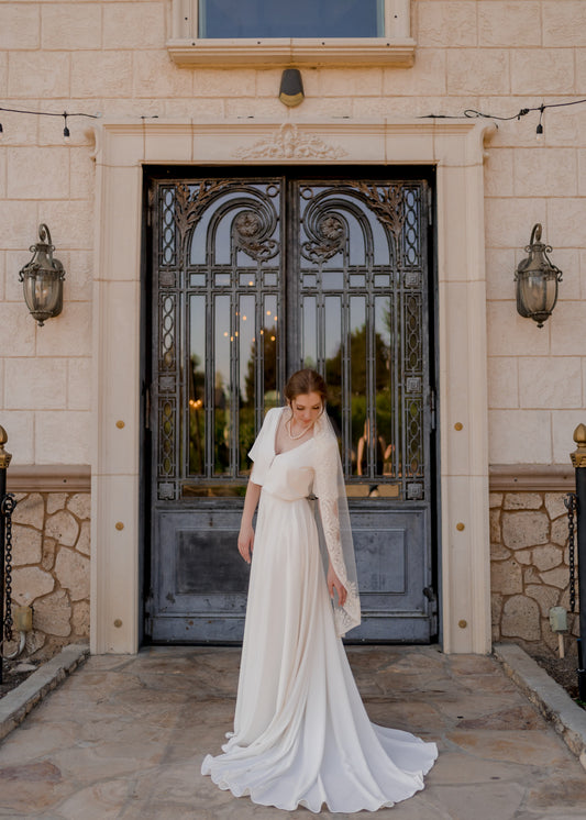 Side view of a bride wearing the Cassie two-piece wedding set, twisting her torso to reveal the softly structured front bodice; the Ireland veil cascades behind her, adding shimmer with its beaded trim.