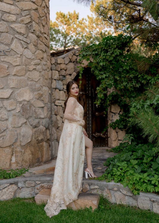 A full length back view of the Reagan gown on a bride as she steps up stone stairs. She twists back and looks at the camera with her leg showing through the wrap skirt. The train and lace sleeves are the focus. 