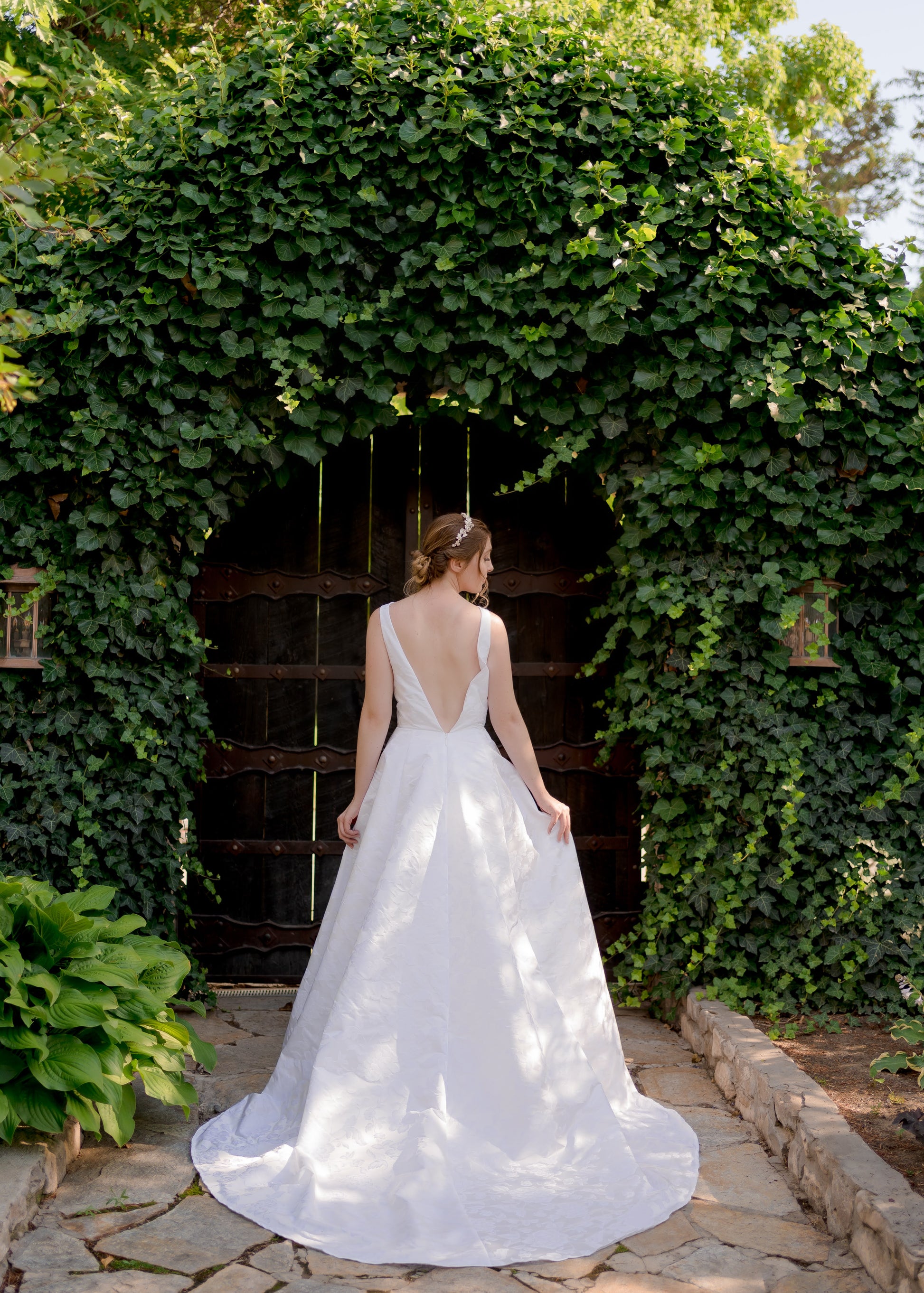 Full back view of a bride standing in a secret garden in the Madison gown, the semi open back dips to the waist and the train extends toward the camera. 