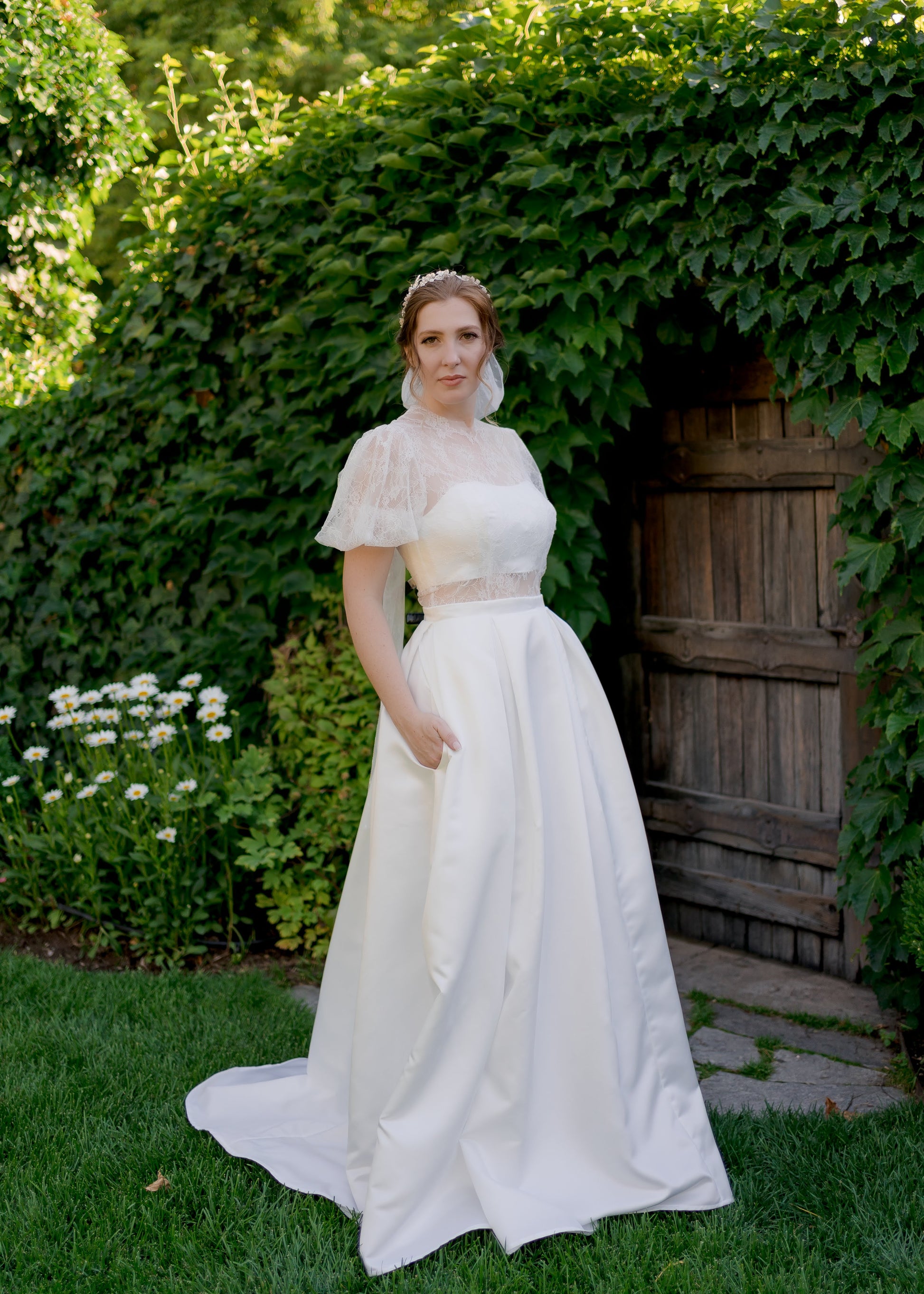 ¾ front view of the Jessa gown in a garden. The structured bando bodice with defined waistband and smooth pleated skirt is highlighted. 