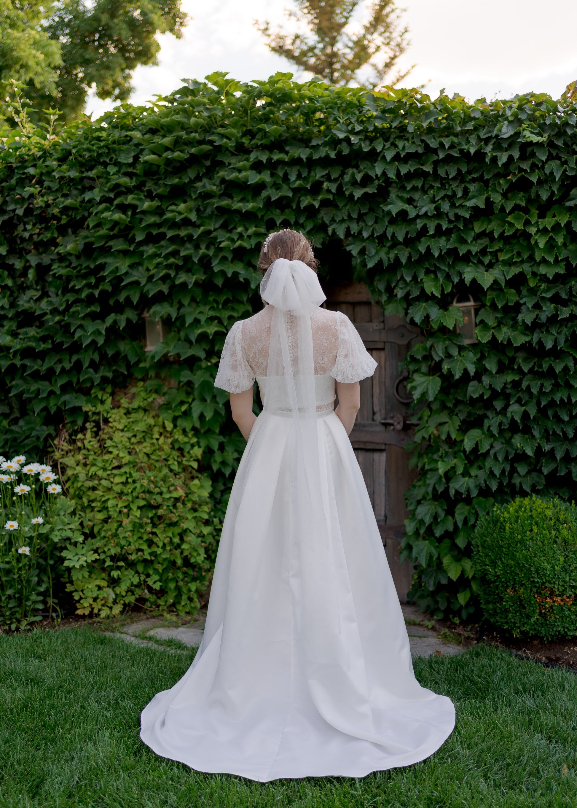 Lace back wedding gown with satin-covered bridal buttons on sheer and bando layers. It has been paired with the  Tess bow veil in Swiss Dot fabric and a fingertip length.