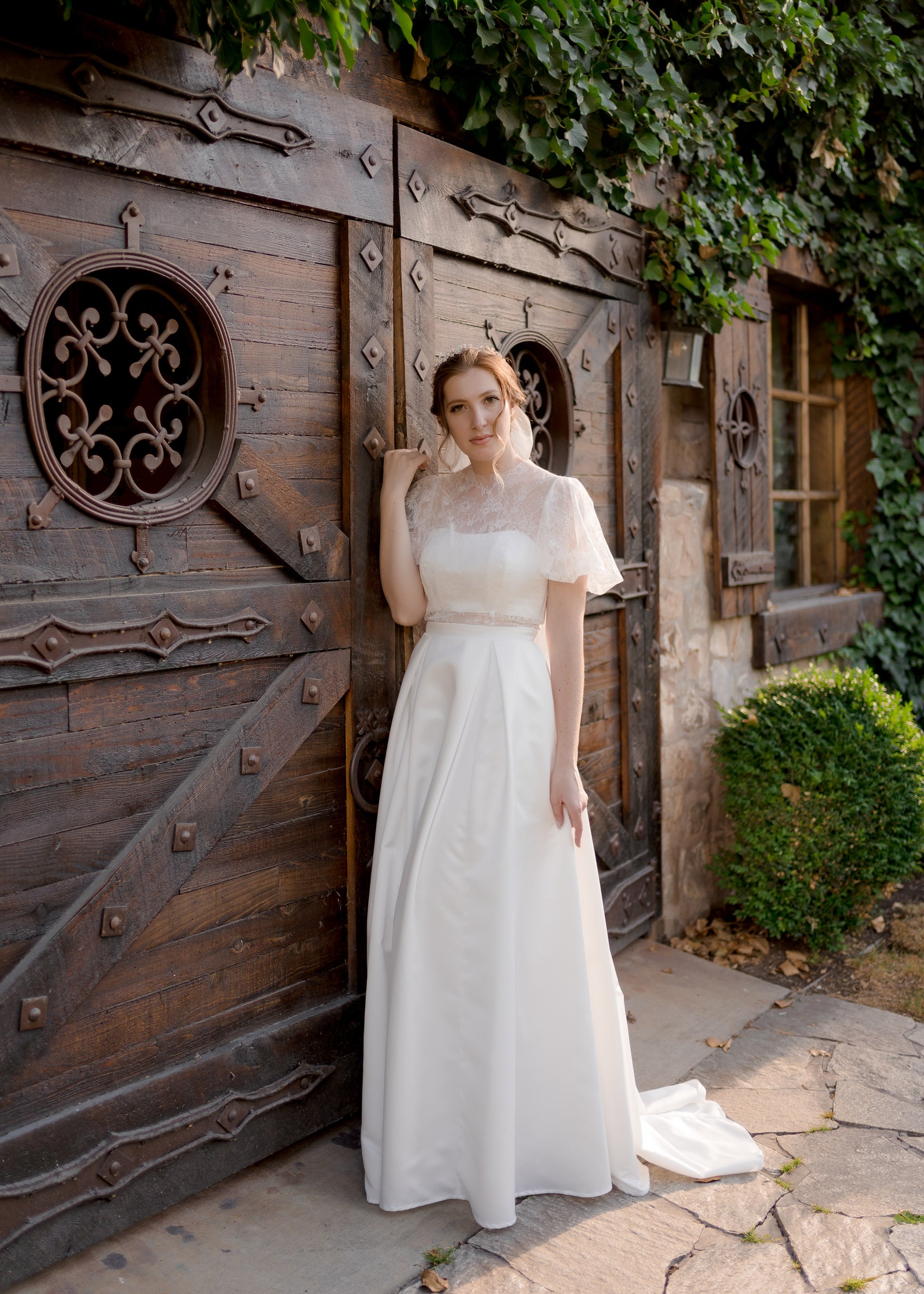 Front full view: Jessa bridal gown with sheer lace bodice, high neckline, and pleated satin skirt.

