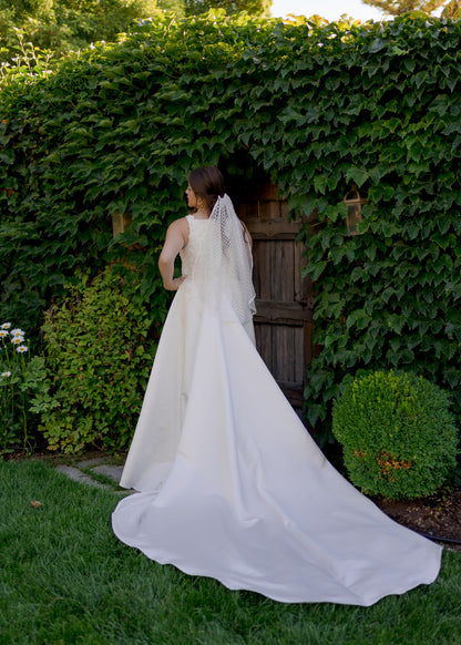 This photo is of a bride in front of an ivy covered wall with a Bride in the sparkly diamond tulle veil, Gloria, and our satin and lace gown, Sabina. The bride faces away from the camera so we can see the fingertip length veil in her hair and the extra long tain extending out on the grass. 