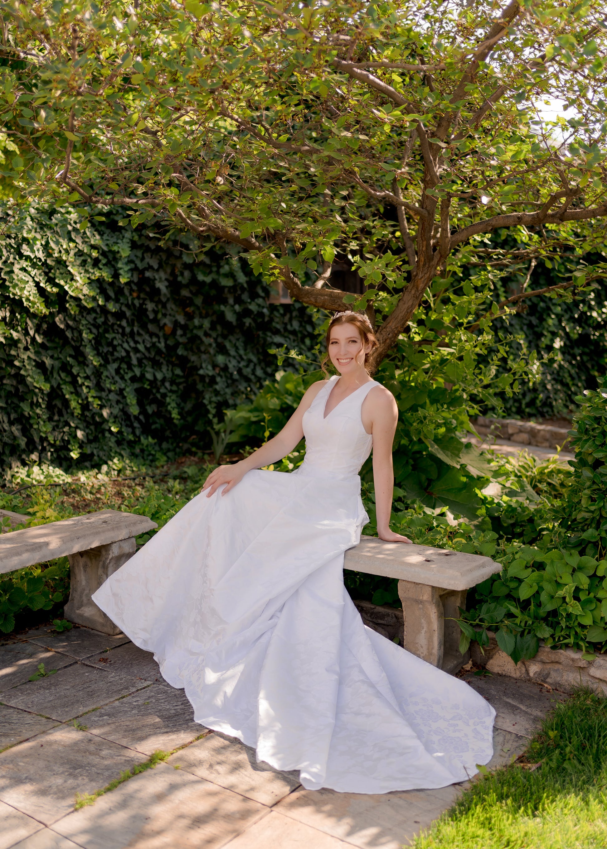 A bride sits on a bench in a garden wearing the madison gown; the flattering and structured bodice flow into the jacquard skirt and 26" long train. 