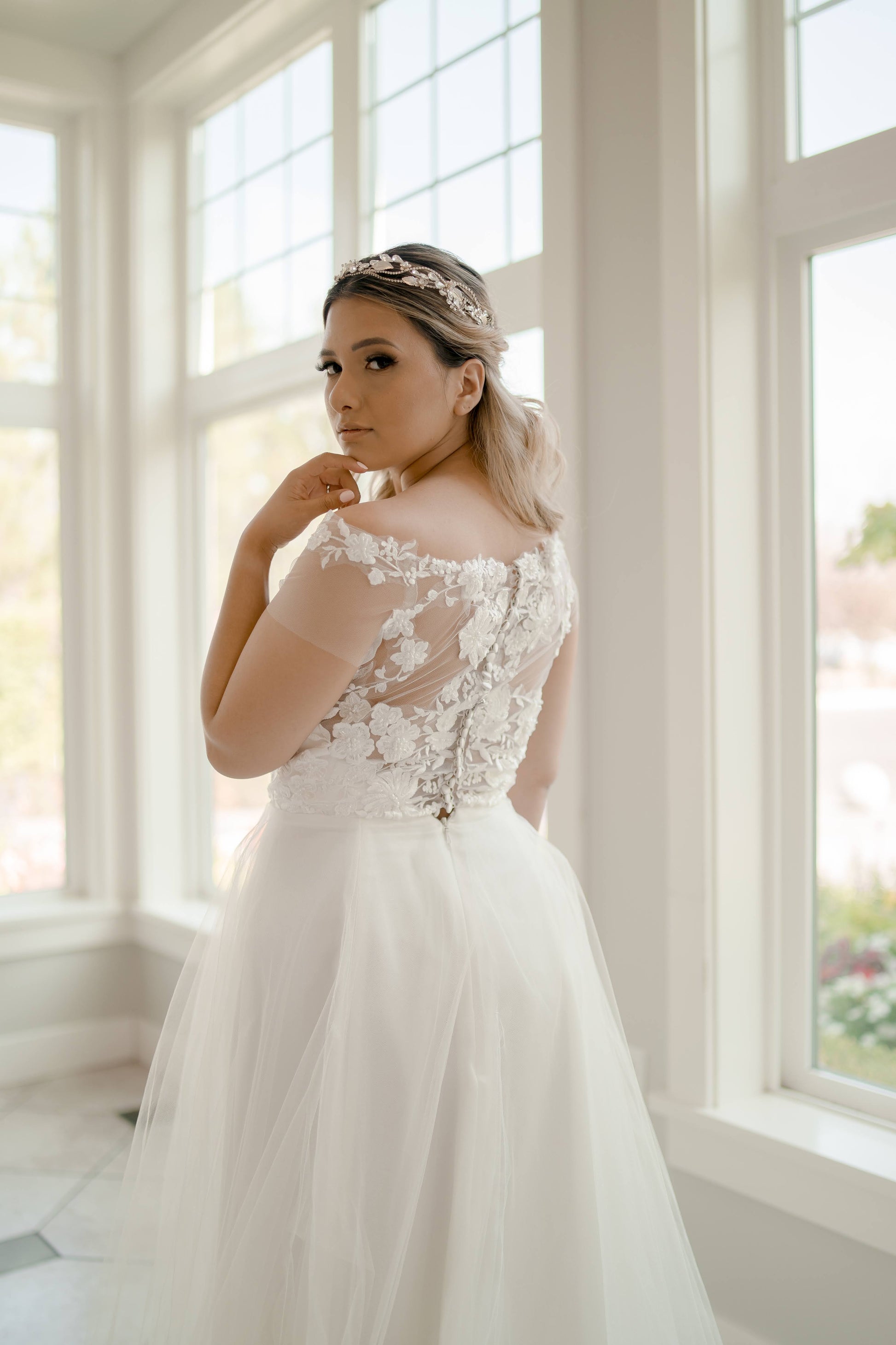 The bride is turned slightly, glancing back over her shoulder toward the camera. The photo highlights the lace and illusion back bodice with satin buttons and shows the airy top of the layered tulle skirt.