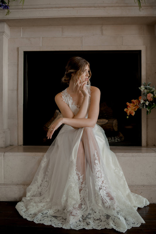 A shot of a model siting in the Marie gown, she sits on a fireplace hearth with her legs crossed and she leans on her knee. The gowns scalloped lace hem and satin slit are the focus. 